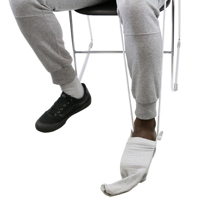 Person wearing gray sweatpants and black sneakers sitting on a chair with one foot in sock aid